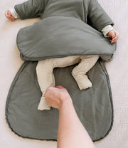 mother changing baby on a white bed. baby is wearing sustainable bamboo convertible footie pajamas in neutral rainbow and unzipped bamboo sleep bag set in sage