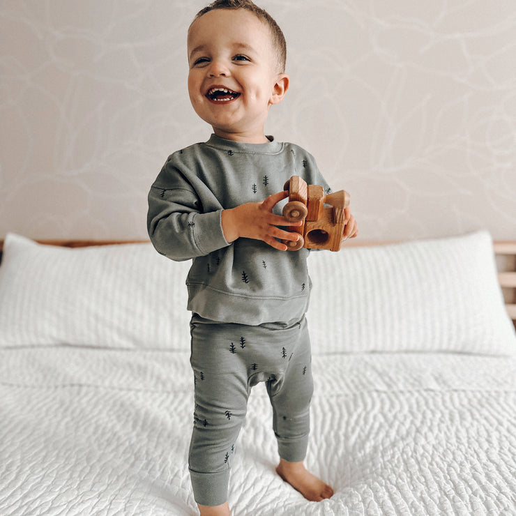 toddler on bed smiling and holding wooden train wearing sustainable baby clothing 2 piece playwear set in forest green with dark green printed trees