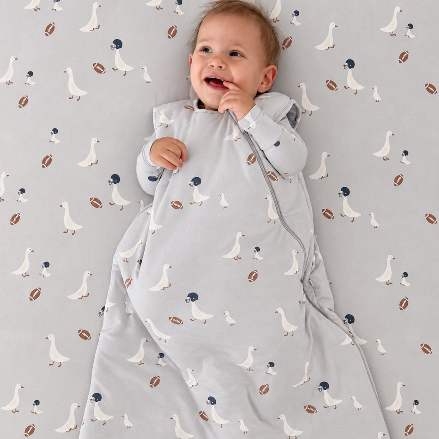 A baby lying on a patterned sheet, wrapped in a light gray zippered swaddle blanket with a white goose print and small brown football accents.