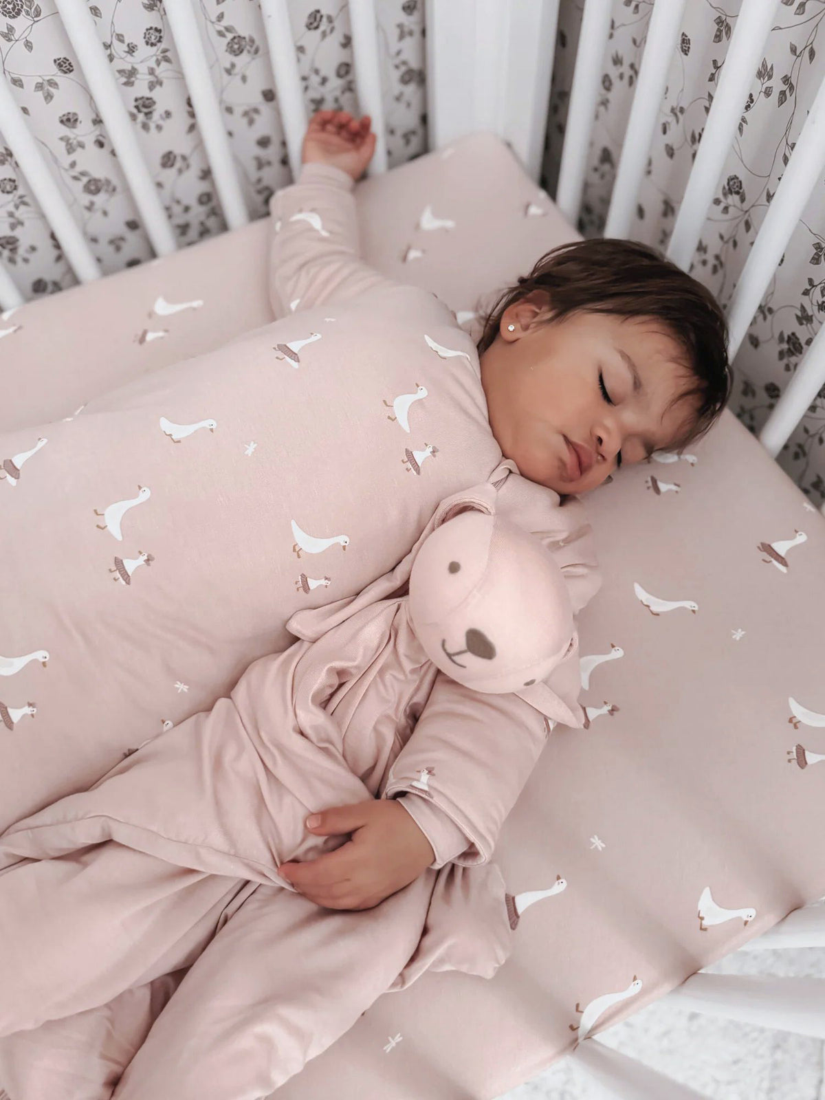 Pink crib sheet with white goose print, baby resting on it with plush bunny