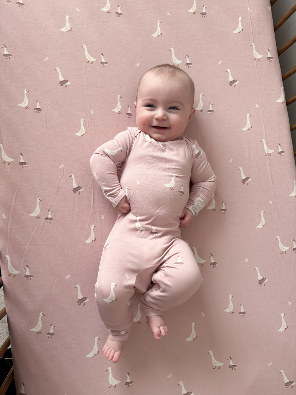 Pink crib sheet with repeating white goose pattern, baby standing on sheet