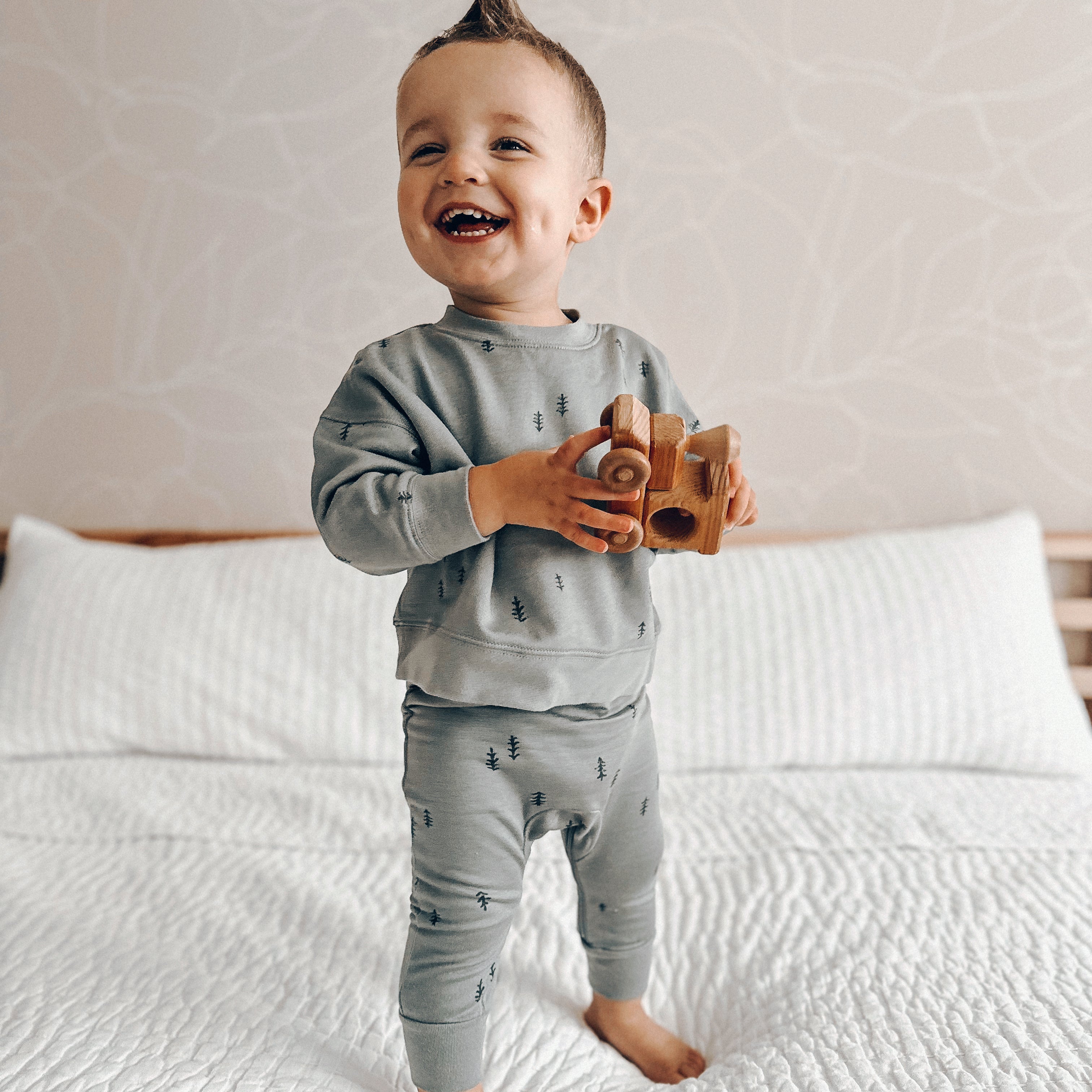 toddler boy standing on white bedsheets, holding a wooden toy train in a fleece jogsuit with diaper zipper