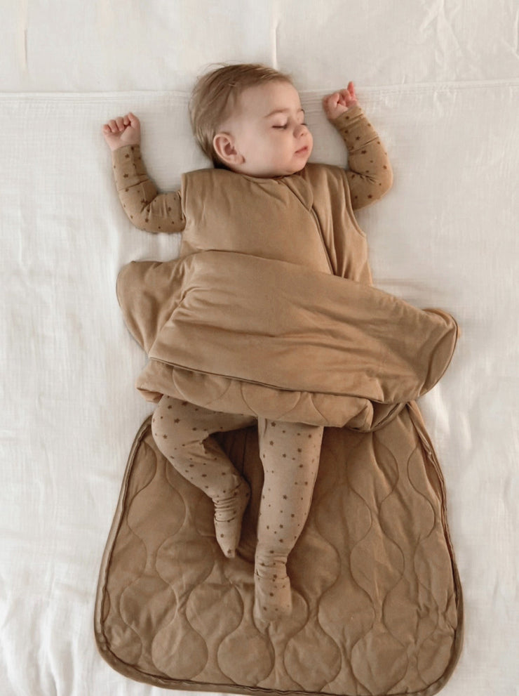baby asleep on a white bed wearing sustainable bamboo convertible footie pajamas and bamboo sleep bag set in brown neutral tones. bag is unzipped for easy diaper change