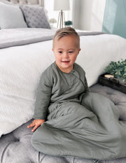 toddler sitting on gray bench at the foot of a white bed wearing a long sleeve toddler sustainable bamboo sleep sack in sage