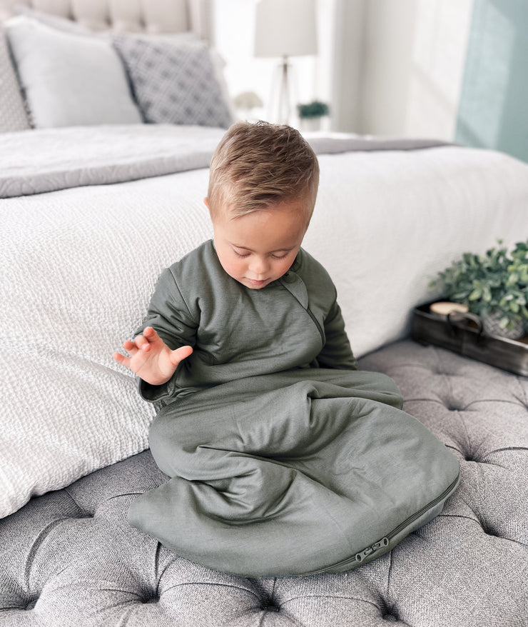 toddler boy sitting on gray bench at the foot of a white bed wearing a long sleeve toddler sustainable bamboo sleep sack in sage