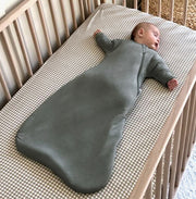 baby asleep on a plaid printed crib sheet in a wooden crib. baby is wearing sustainable transitional swaddle sleep bag in muted green color