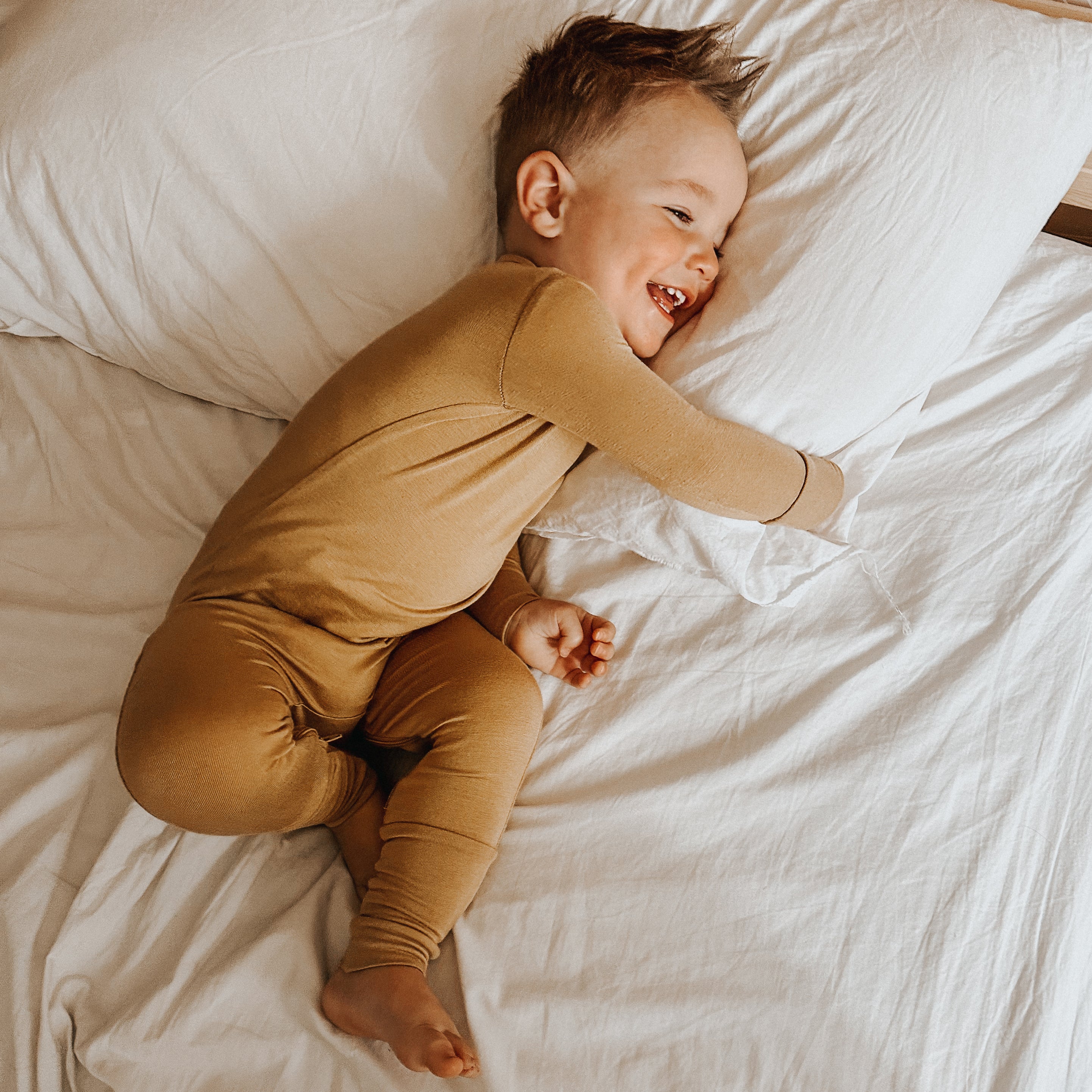 toddler boy smiling and hugging a white pillow on a white bedsheet wearing convertible pajamas with diaper zipper for easy diaper changes