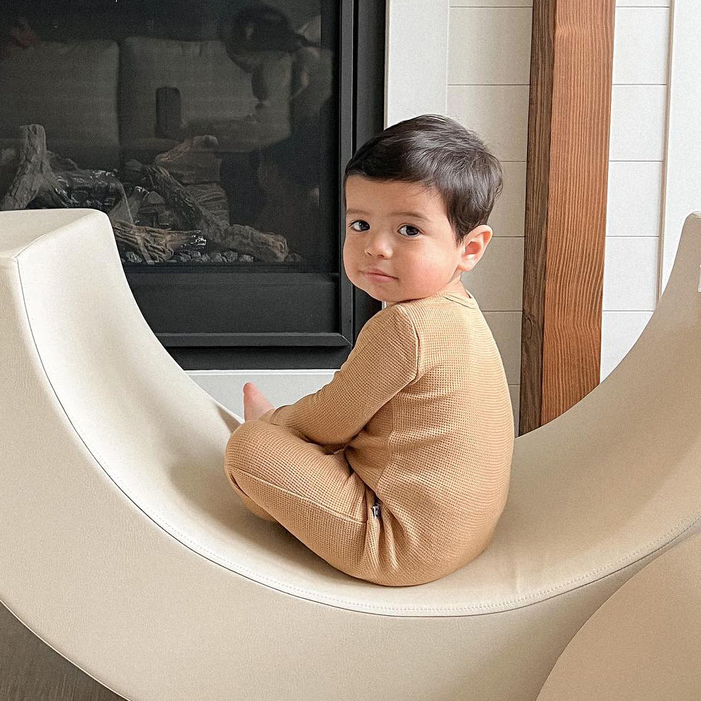 baby boy sitting on a beige cushion in front of a fireplace with cappuccino colored waffle convertible pajamas, looking back at the camera