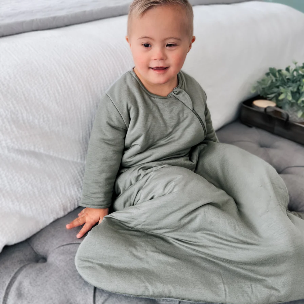 toddler boy sitting on a bench in a sage colored long sleeve sleep bag