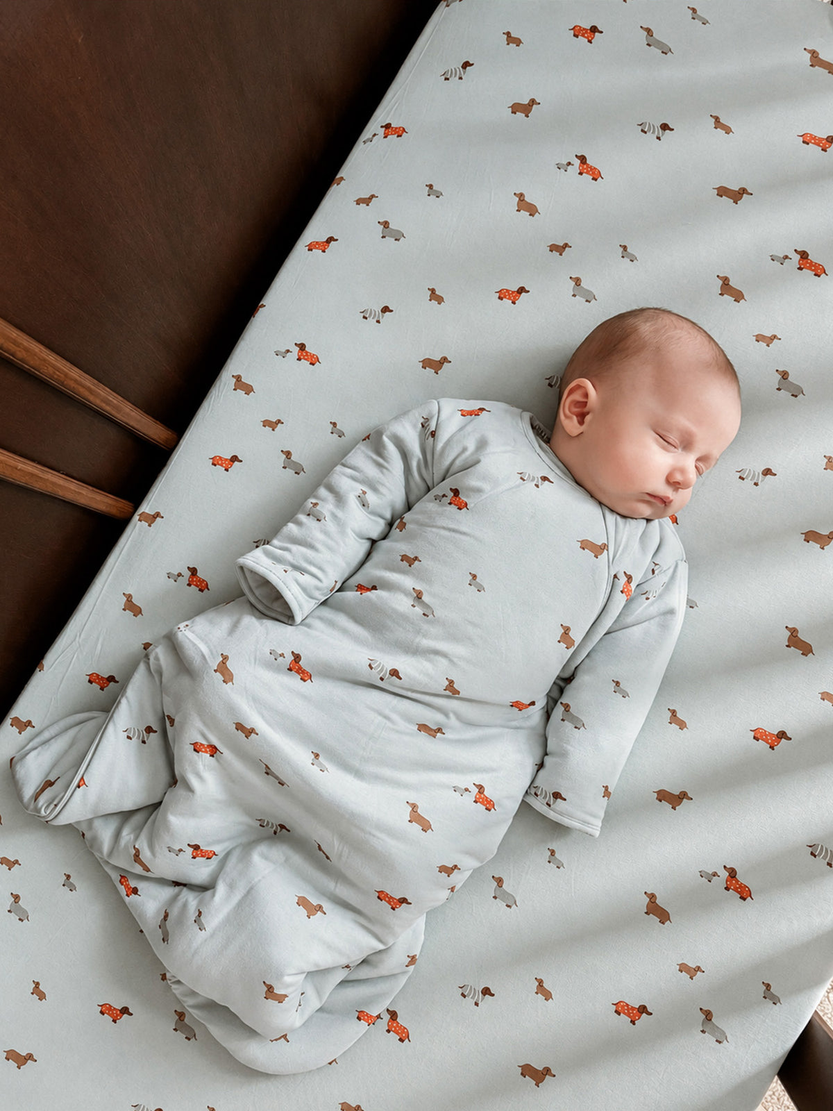 Light blue crib sheet corner with orange dog print pattern, baby lying on it