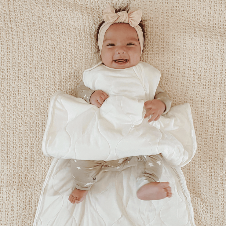 baby girl on a light beige sheet wearing undyed hypoallergenic sleep bag made for sensitive skin paired with neutral colored convertible footie pajamas with subtle star print. sleep sack is unzipper during a diaper change
